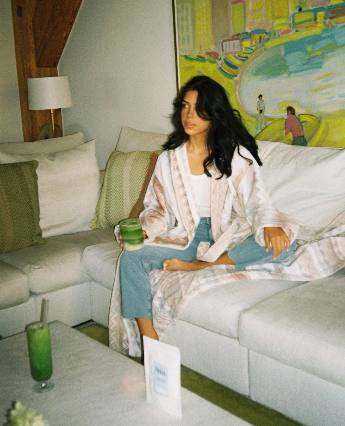 A woman sits on a white sectional sofa in a cozy living room setting, wearing a white patterned kimono robe over a white top and light blue jeans. A green matcha latte drink is held in her hand, with a colorful painting and wooden ceiling beams in the background.